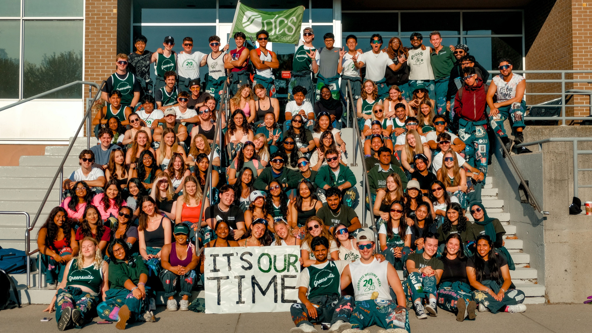A large group of university students packed in rows of stone stairs leading to a building for a group photo. They're all smiling with DCS banners and a poster reading, 'It's our time.'