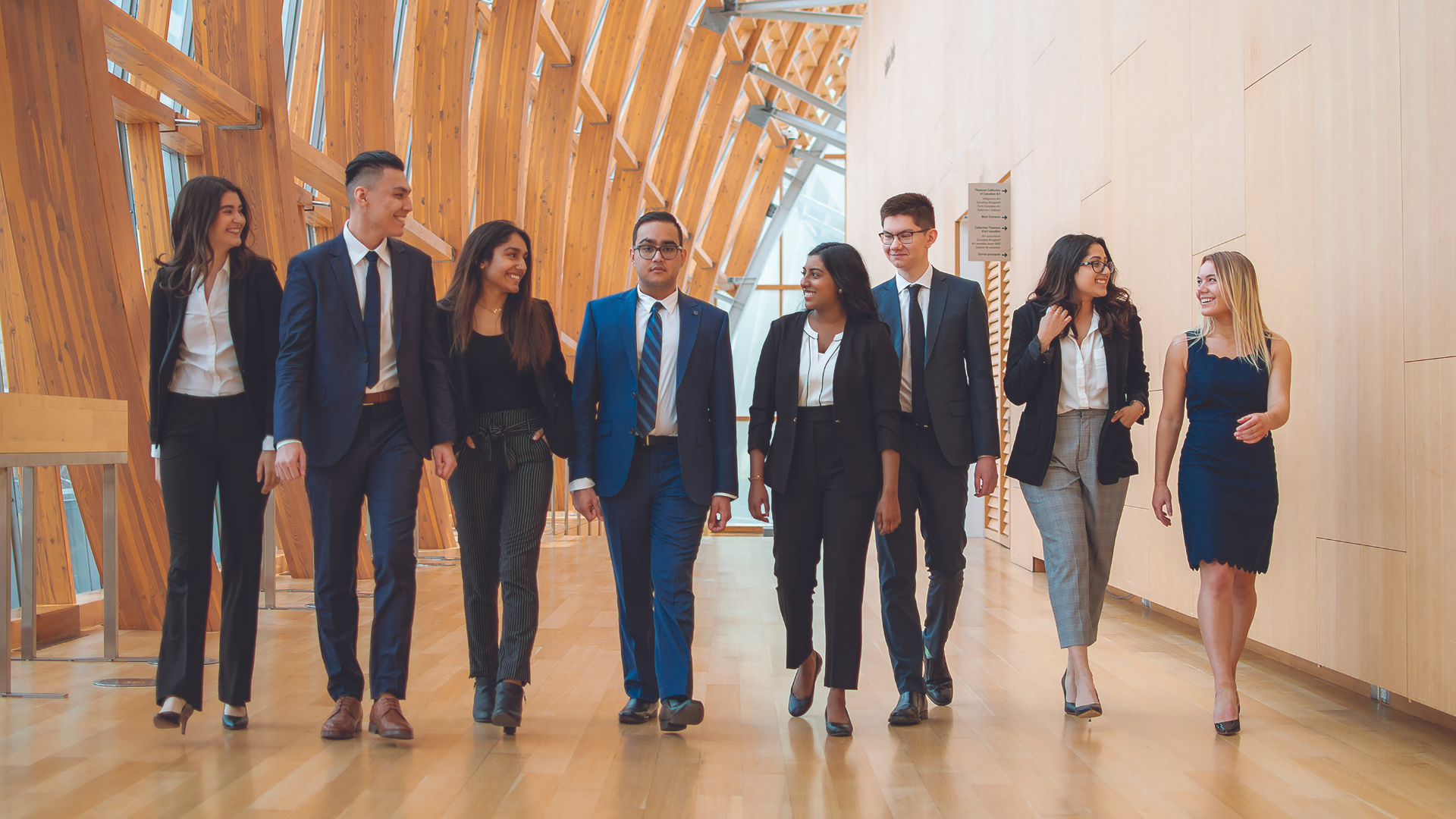 Eight young businesspeople (five women and three men) walking down a cream-finished office hallway smiling and talking.