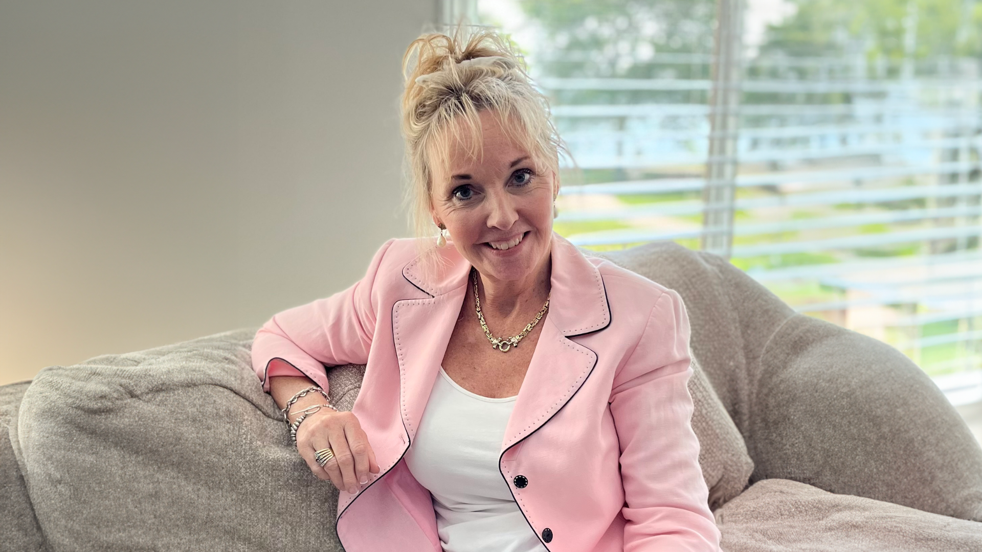 Teal McAteer, a smiling White woman wearing a pink jacket and white shirt, leaning on a couch.